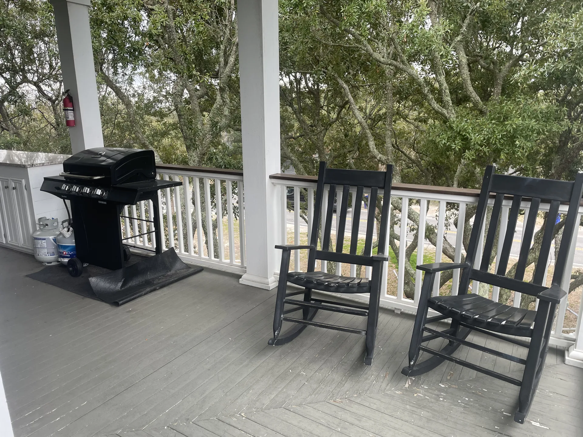 Covered wraparound porch with black rocking chairs, gas grill, and live oak views