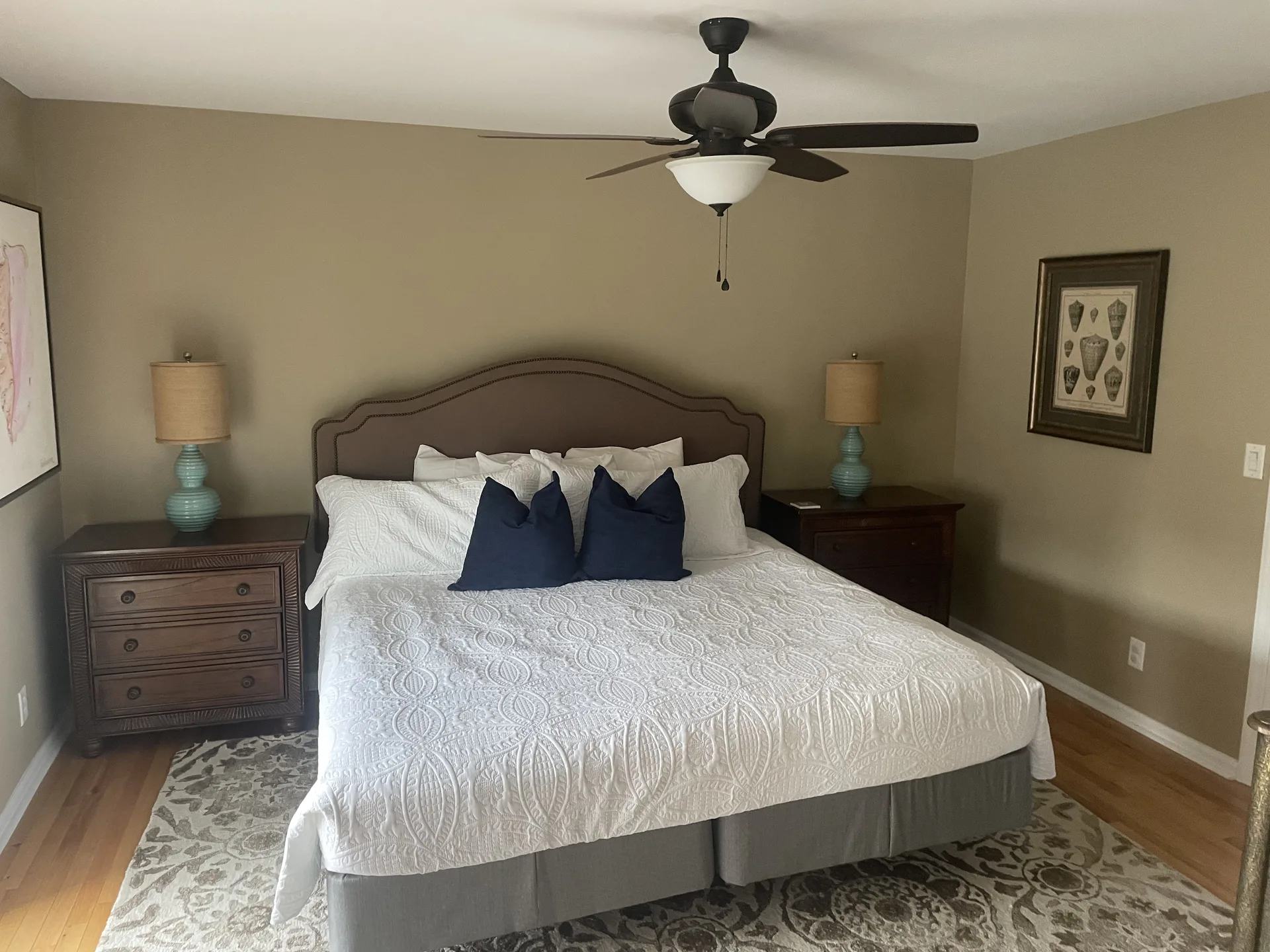 King bedroom with navy accent pillows, ceiling fan, and coastal shell artwork